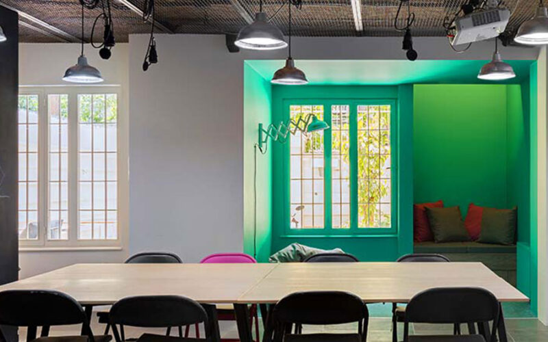 Workshop interior with a long wooden table and black chairs, a bright green painted alcove with a window seat and industrial pendant lamps