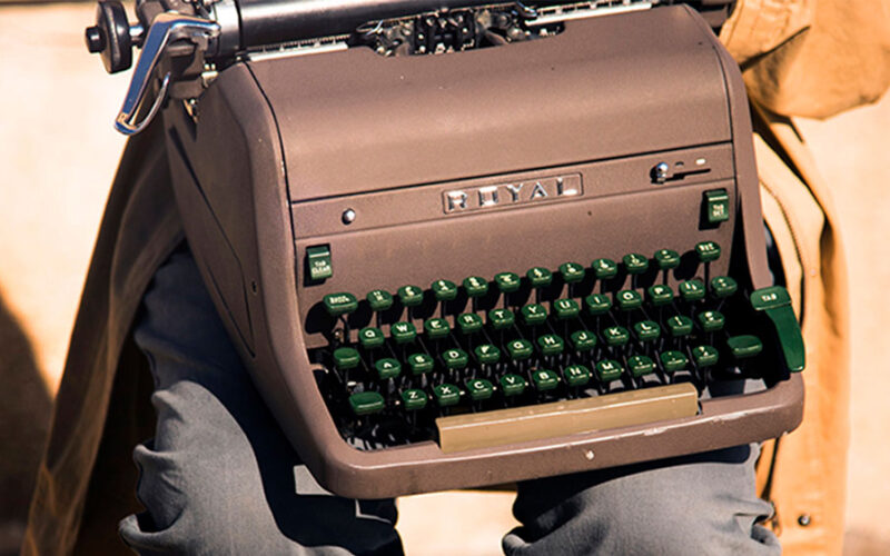 A person in a beige coat carrying a brown Royal manual typewriter with green keys, photographed in warm evening light