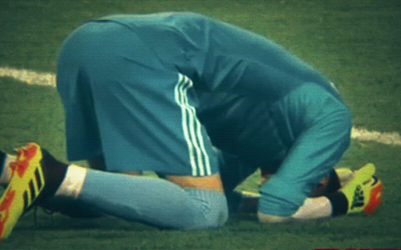 Grainy video still of a footballer in a blue kit kneeling face-down on the grass, filmed with a VHS-style scan-line effect
