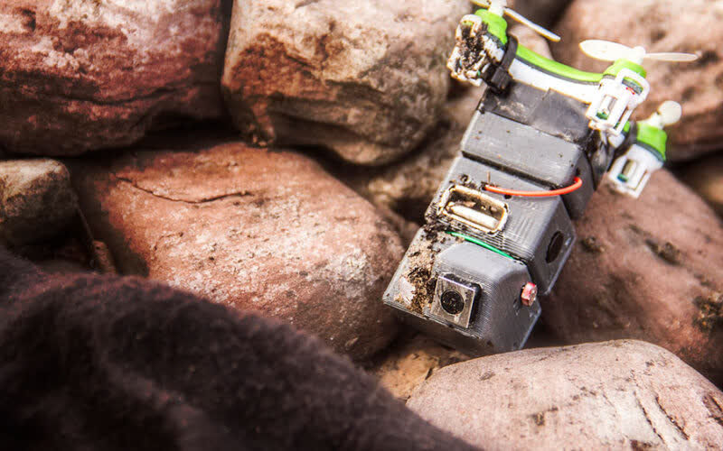 A small hand-built quadcopter with exposed circuit board, wiring and a camera module resting on a pile of rust-coloured rocks