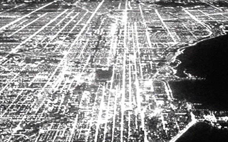 Black-and-white aerial night view of a city grid, with streets traced as bright lines of light
