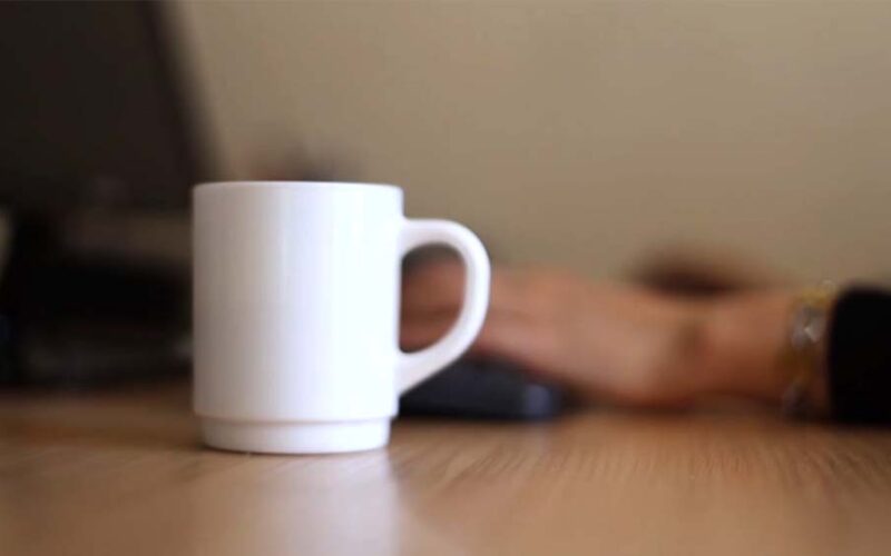 Close-up of a white ceramic mug on a wooden desk with a person's hands typing on a keyboard behind it